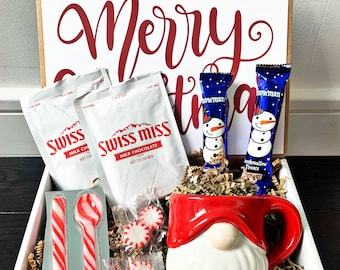 May include: A white gift box with a red "Merry Christmas" sign on top. The box contains a white gnome mug, two red and white striped spoons, three packs of peppermint candies, two packets of Swiss Miss hot cocoa mix, and two snowman-shaped chocolate bars.
