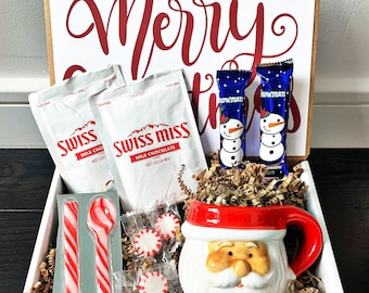 May include: A gift box with a festive theme, containing hot cocoa mix, peppermint candies, candy spoons, and snowman-themed chocolate bars. A Santa-faced mug with a red handle and trim is also included. The box features a sign that says "Merry Christmas" in red script.