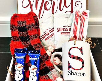 May include: A white gift box filled with a red and black plaid fuzzy sock, two blue snowman-shaped marshmallow treats, two packets of Swiss Miss hot cocoa mix, a white mug with a red and black plaid letter "S" and the name "Sharon" printed on it, two candy canes, and a white spoon.