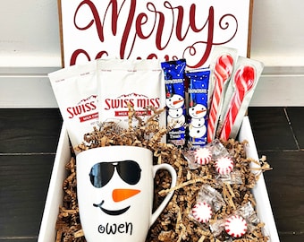 May include: White gift box with the words "Merry Christmas" in red lettering. The box contains a white mug with a snowman face, two packages of Swiss Miss hot chocolate mix, two snowman-shaped chocolate bars, candy canes, and peppermint candies.