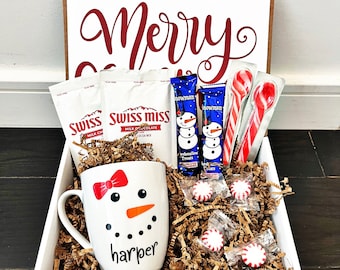 May include: A white gift box with the words "Merry Christmas" on the lid. The box is filled with a snowman mug, hot chocolate mix, candy canes, and peppermint candies. The mug has a snowman design and the name "harper" written on it.