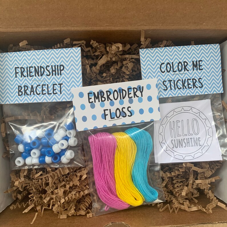 May include: A craft kit with three items: a friendship bracelet kit with blue and white beads, embroidery floss in pink, yellow, and blue, and a coloring page with the text "Hello Sunshine".