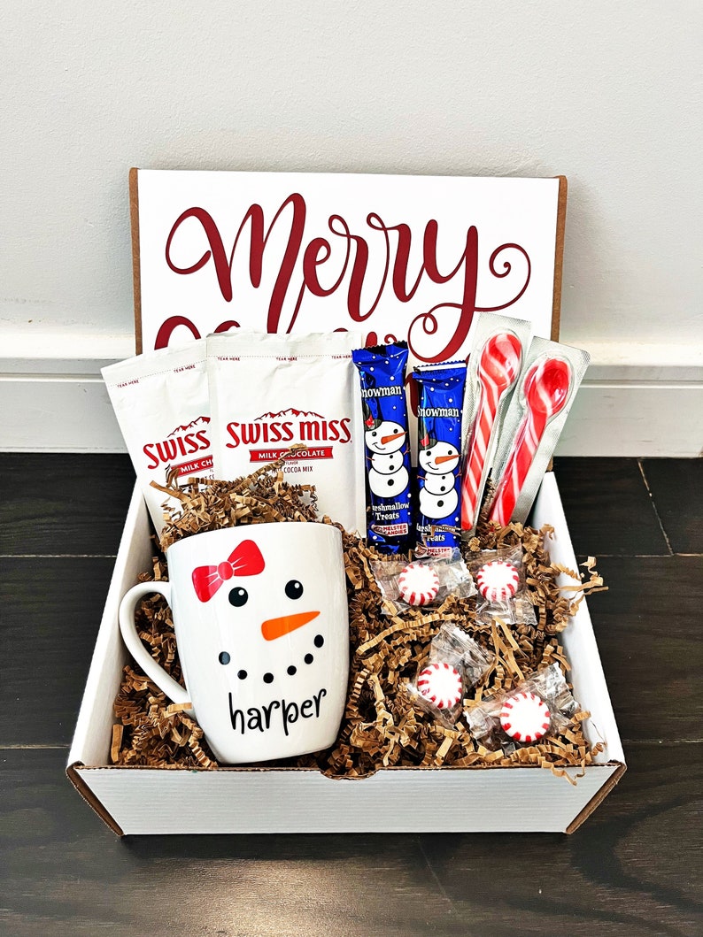 May include: A white gift box with the words "Merry Christmas" on the lid. The box is filled with a snowman mug, hot chocolate mix, candy canes, and peppermint candies. The mug has a snowman design and the name "harper" written on it.