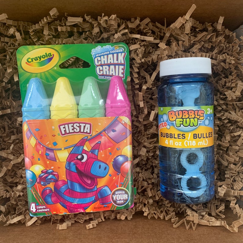 May include: A box filled with brown packing paper contains a package of four colorful sidewalk chalk sticks labeled "Crayola Fiesta Washable Sidewalk Chalk Craie" and a blue bottle of bubble solution labeled "Bubbles Fun 3+ Bubbles/Bulles 4 fl oz (118 mL)"