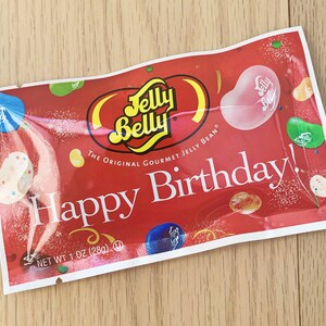 Może przedstawiać: Czerwono-biały woreczek z cukierkami Jelly Belly z napisem "Happy Birthday!" i "The Original Gourmet Jelly Bean". Woreczek jest ozdobiony kolorowymi żelkami i r&oacute;żową fasolką Jelly Belly z logo.