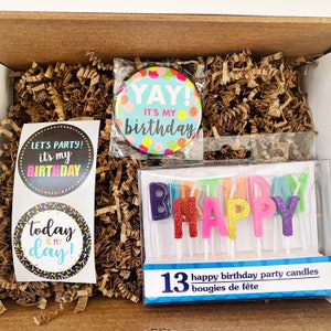 Może przedstawiać: Pudełko prezentowe na przyjęcie urodzinowe z niebieskim i białym guzikiem z napisem "YAY! It's my birthday", dwie naklejki z napisami "Let's party! It's my birthday" i "Today is my day!" oraz opakowanie 13 kolorowych świeczek, kt&oacute;re tworzą napis "HAPPY".