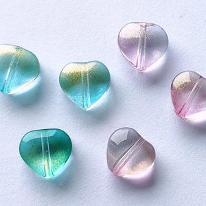 May include: Six heart-shaped glass beads in shades of teal, green, and pink. Each bead has a gradient effect with a shimmering gold or silver accent. The beads are translucent and have a hole for stringing.