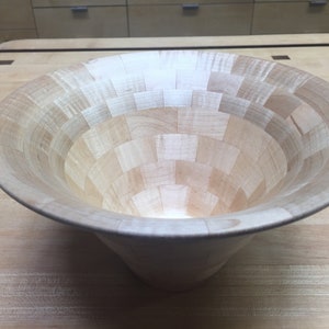 May include: A large, wooden bowl made of maple wood. The bowl is made of many small pieces of wood that are arranged in a geometric pattern. The bowl is light brown in color and has a smooth, polished finish.