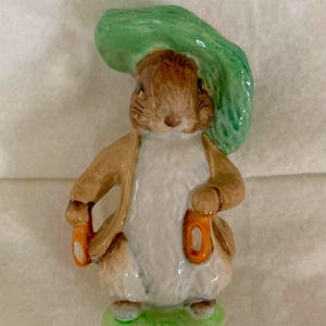 May include: A ceramic figurine of a rabbit, wearing a tan jacket and holding two orange objects. The rabbit has a white chest and a green hat. The figurine is standing on a green base.