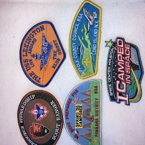 May include: A collection of embroidered patches with various designs. One patch features a ship with text "USS Lexington". Another shows a map of Long Island, NY. Other patches include text "I Camped in Space" and "Scout Ranger".