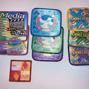 May include: A collection of Girl Scout badges featuring various themes, including media, connections, and citizenship. The badges are colorful and feature different designs, such as a film reel, puzzle pieces, and a dove.
