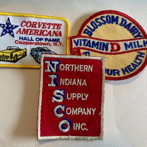May include: Three embroidered patches. The first patch is yellow with a red, white, and blue star and the text "Corvette Americana Hall of Fame Cooperstown, N.Y.". The second patch is red with white text that reads "Northern Indiana Supply Company Inc.". The third patch is yellow with blue and red text that reads "Blossom Dairy Vitamin D Milk Our Health".