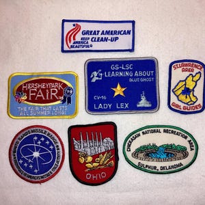 May include: A collection of eight embroidered patches. The patches feature various designs, including a blue and white patch with the text "Hersheypark Fair", a red, white, and blue patch with the text "Great American Clean-Up", a blue patch with the text "GS-LSC Learning About Blue Ghost", a white patch with the text "St. Lawrence Area Girl Guides", a red patch with the text "Ohio", a blue and white patch with the text "White Sands Missile Range", and a white patch with the text "Chickasaw National Recreation Area".