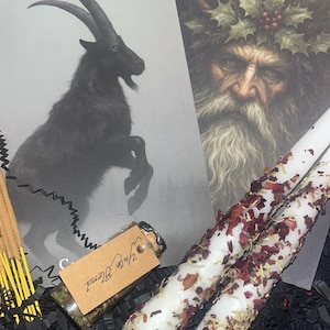 May include: A collection of esoteric items, featuring two white candles decorated with dried rose petals and herbs. A black and white illustration of a goat and a portrait of a bearded man with a holly wreath are also visible. A small glass vial labelled "Yule Blend" is present.