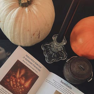 May include: A still life arrangement featuring a white pumpkin, an orange pumpkin, and a black candle in a glass holder. An open book with the title "Hallowed Harvest" and a small cauldron are also present. The theme is related to Halloween.