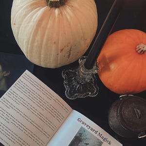 May include: A still-life composition featuring two pumpkins, one white and one orange, with a lit black candle in a glass holder. An open book with the title "Graveyard Magick" and a small cauldron complete the Halloween theme.