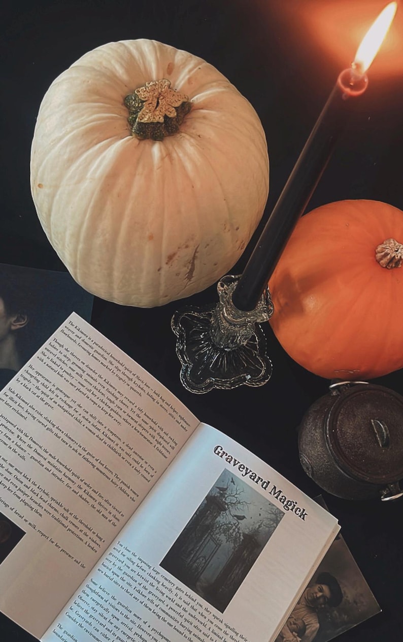 May include: A still-life composition featuring two pumpkins, one white and one orange, with a lit black candle in a glass holder. An open book with the title "Graveyard Magick" and a small cauldron complete the Halloween theme.