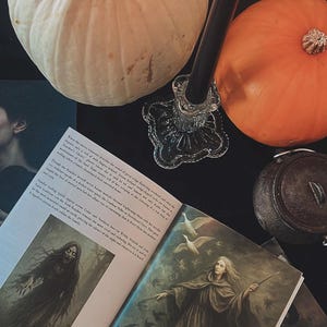 May include: A still life featuring a white pumpkin, an orange pumpkin, a lit black candle in a glass holder, and an open book with illustrations. The book's title is "Nicneven". The scene is set on a dark surface.