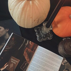 May include: A still life arrangement featuring two pumpkins, one white and one orange, with a lit black candle in a glass holder. An open book titled "SHADOW WORK" and other mystical items are arranged on a black surface.