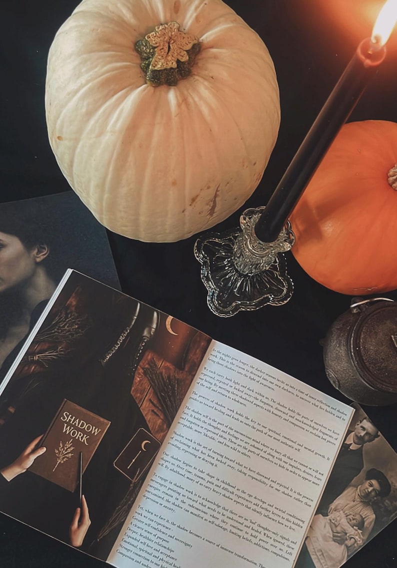 May include: A still life arrangement featuring two pumpkins, one white and one orange, with a lit black candle in a glass holder. An open book titled "SHADOW WORK" and other mystical items are arranged on a black surface.