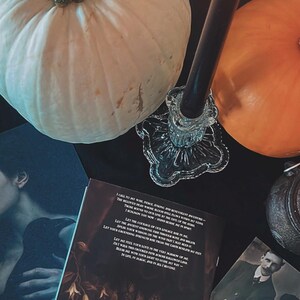 May include: A still-life arrangement featuring a white pumpkin, an orange pumpkin, a lit black candle in a glass holder, and vintage photographs. A book with gothic illustrations and text is also present, creating a Halloween or autumn theme.