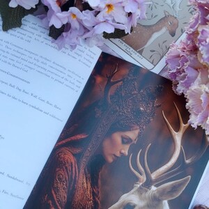 May include: An open book with a detailed illustration of a woman with a deer. The woman is wearing a crown and a red dress. A tarot card with a deer illustration is visible. Pink flowers are in the background.