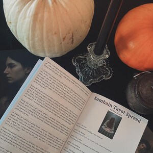 May include: A still-life arrangement featuring a white pumpkin, an orange pumpkin, a lit black candle in a glass holder, and an open book titled "Samhain Tarot Spread." The scene is set on a dark surface, evoking a mystical theme.