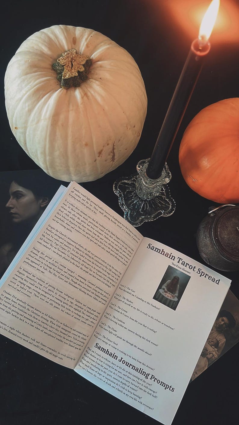 May include: A still-life arrangement featuring a white pumpkin, an orange pumpkin, a lit black candle in a glass holder, and an open book titled "Samhain Tarot Spread." The scene is set on a dark surface, evoking a mystical theme.