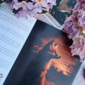 May include: An open book with a detailed illustration of a fox against a sunset backdrop. A tarot card with a fox and ivy design is visible. Pink flowers and text are in the image.