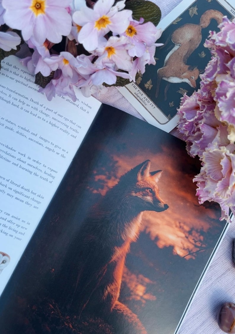 May include: An open book with a detailed illustration of a fox against a sunset backdrop. A tarot card with a fox and ivy design is visible. Pink flowers and text are in the image.
