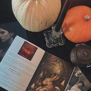 May include: A still-life arrangement featuring a white pumpkin, an orange pumpkin, a lit black candle in a glass holder, and an open book with the text "Magick Mushrooms". Other items include a small cauldron and vintage photographs.