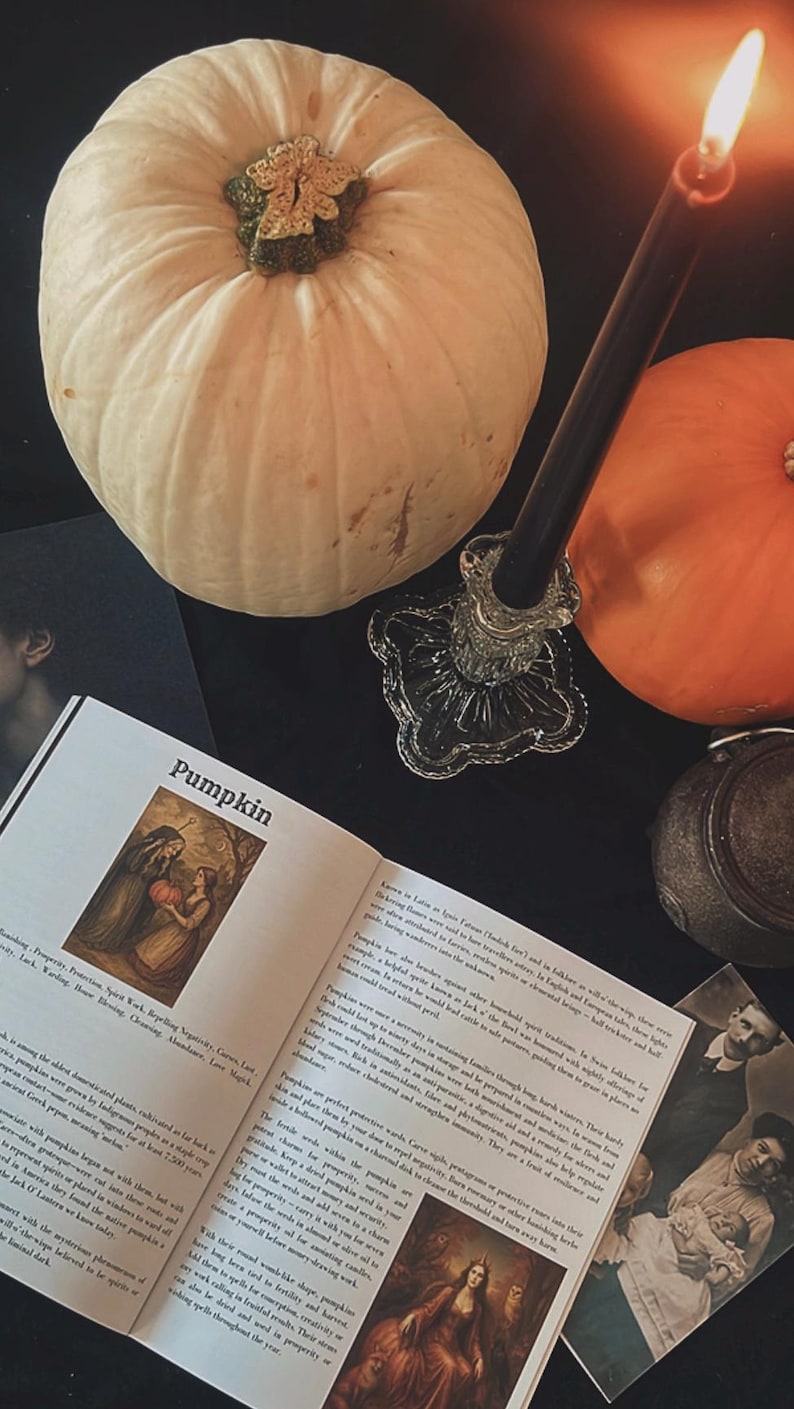 May include: A still life featuring a white pumpkin, a lit black candle in a glass holder, and an open book with the title "Pumpkin." The book displays illustrations and text. A small orange pumpkin and a vintage photo are also present.