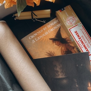 May include: A gift box containing sunflowers, candles, a key, a rolled print, a book titled "Crowned by Sunlight A Lammas Grimoir," and incense. The box is lined with black tissue paper, creating a mystical theme.
