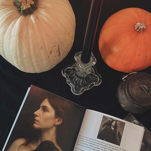 May include: A still-life arrangement featuring a white pumpkin, an orange pumpkin, a lit black candle in a glass holder, and an open book titled "The Morrigan." The book displays an illustration of a woman with a raven.