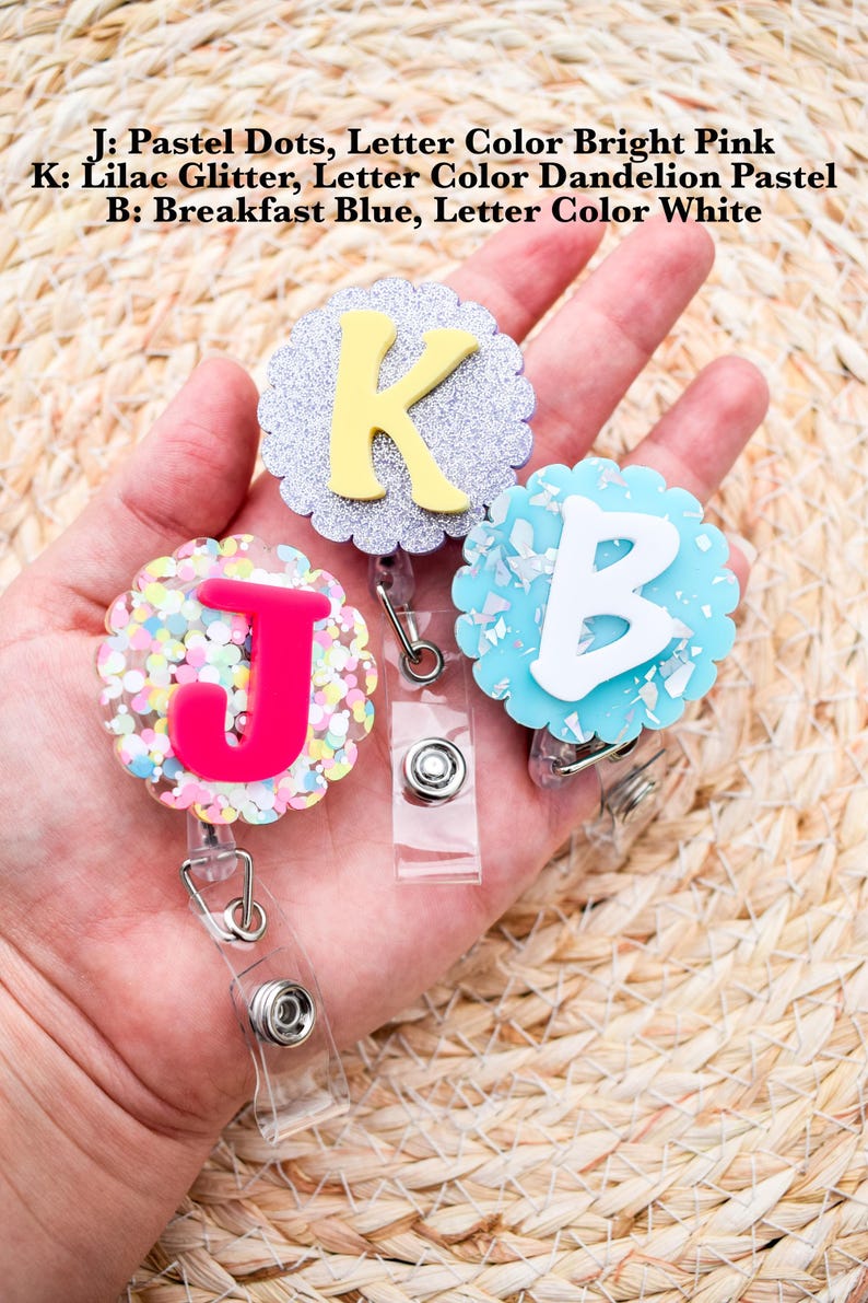 May include: Three badge reels with the letters J, K, and B. The J reel has pastel dots and a bright pink letter. The K reel has lilac glitter and a dandelion pastel letter. The B reel has breakfast blue and a white letter. The text on the image reads: J: Pastel Dots, Letter Color Bright Pink, K: Lilac Glitter, Letter Color Dandelion Pastel, B: Breakfast Blue, Letter Color White.