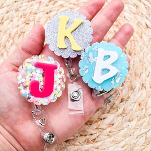 May include: Three decorative badge reels with scalloped edges, each featuring a different letter: a yellow "K" on silver glitter, a white "B" on blue, and a pink "J" with colorful confetti. Each reel has a clear plastic strap and a metal clip.