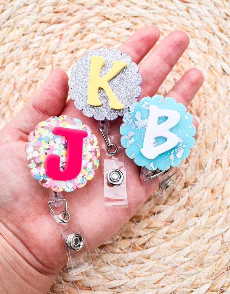 May include: Three decorative badge reels with scalloped edges, each featuring a different letter: a yellow "K" on silver glitter, a white "B" on blue, and a pink "J" with colorful confetti. Each reel has a clear plastic strap and a metal clip.