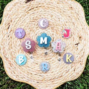 May include: A collection of personalized badge reels with glittery letters and floral designs. The letters are in various colors, including pink, blue, and purple. The badge reels are arranged on a woven, straw-like surface.