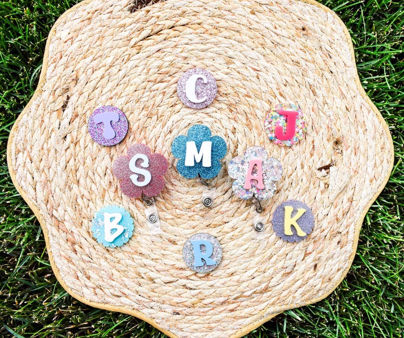May include: A collection of personalized badge reels with glittery letters and floral designs. The letters are in various colors, including pink, blue, and purple. The badge reels are arranged on a woven, straw-like surface.