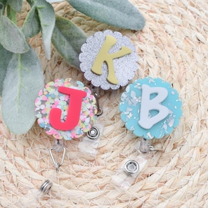 May include: Three badge reels with scalloped edges, each featuring a different letter: a yellow "K" on a glittery silver background, a red "J" with colorful confetti, and a white "B" on a blue background. The reels have a clear plastic retractable cord.