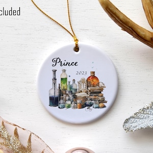 May include: White ceramic ornament with a gold string, featuring a watercolor illustration of potion bottles and books. The text "Prince 2023" is written on the ornament.