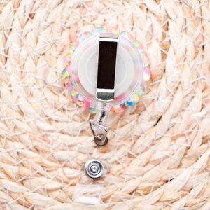 May include: A clear, round retractable badge reel with a silver metal clip and a white center. The reel is decorated with colorful sequins and sits on a woven, light brown surface. The badge reel has a clear plastic strap and a silver metal snap.