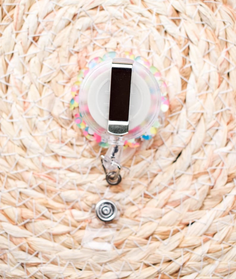 May include: A clear, round retractable badge reel with a silver metal clip and a white center. The reel is decorated with colorful sequins and sits on a woven, light brown surface. The badge reel has a clear plastic strap and a silver metal snap.