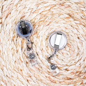 May include: Two clear plastic retractable badge holders with metal clips and black accents. The badge reels are on a woven, light brown surface.