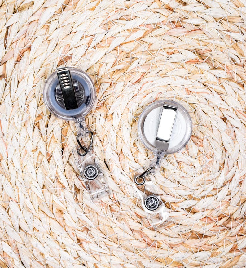 May include: Two clear plastic retractable badge holders with metal clips and black accents. The badge reels are on a woven, light brown surface.