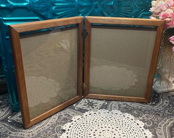 May include: A wooden double picture frame with a light brown wood grain finish. The frame has two openings for pictures and a hinged design that allows the frame to be opened and closed.