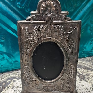 May include: A rectangular silver-toned metal picture frame featuring an oval center opening. The frame is decorated with embossed floral and grape vine motifs, a prominent crest at the top, and four small feet at the base for stability.