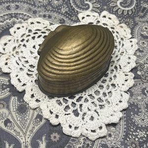 May include: A vintage brass clam shell-shaped trinket box with a hinged lid. The box is resting on a white, crocheted doily, set against a patterned fabric background. The box has a textured, ridged surface and a warm, metallic finish.