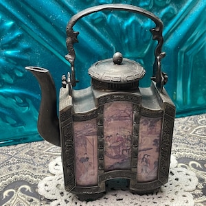 May include: Antique silver-toned teapot with a decorative handle and spout. The body features panels with Asian-inspired artwork. The teapot has a lid with a small knob and sits on a decorative base. The overall design is ornate and detailed.