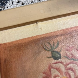 Puede incluir: Primer plano de una pintura enmarcada de una rosa rosa con un insecto verde en el tallo. La pintura est&aacute; sobre lienzo con un fondo marr&oacute;n.